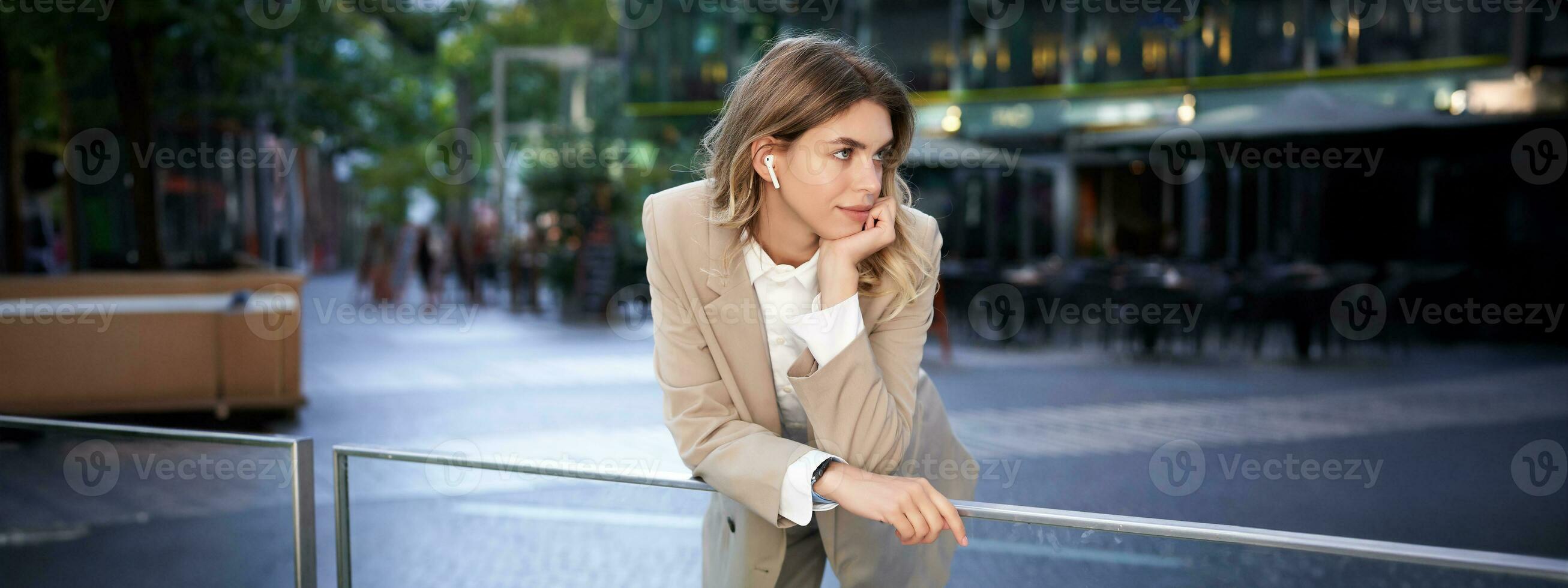 elegante corporativo mujer en inalámbrico auriculares y traje, posando en calle cerca oficina edificios foto