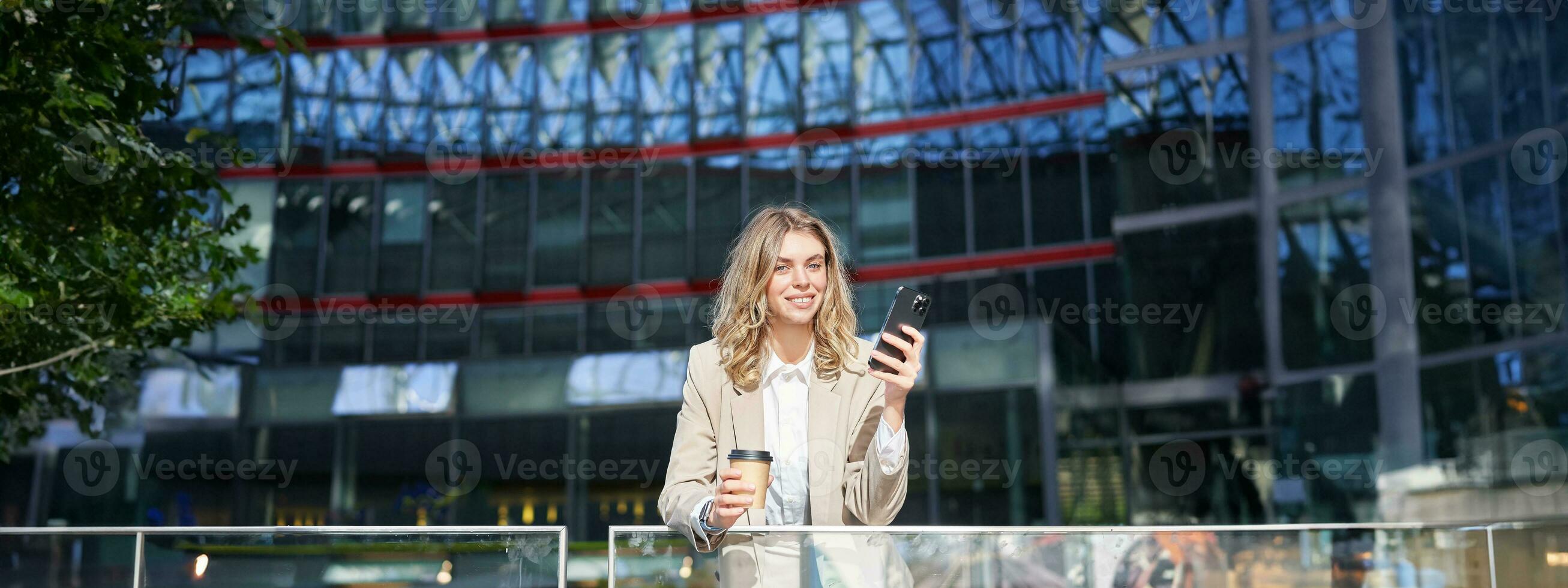 Businesswoman in suit standing with smartphone on street, drinking coffee, using mobile phone app, browsing internet, having day break photo