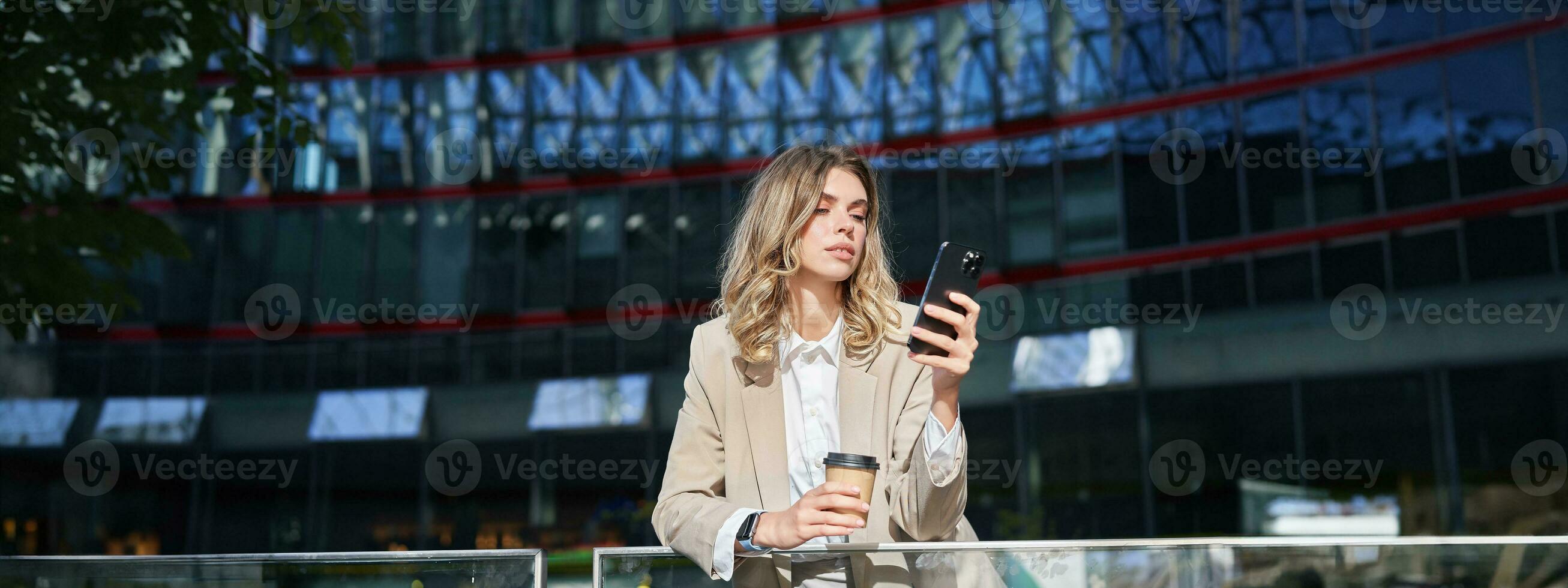 Businesswoman in suit standing with smartphone on street, drinking coffee, using mobile phone app, browsing internet, having day break photo