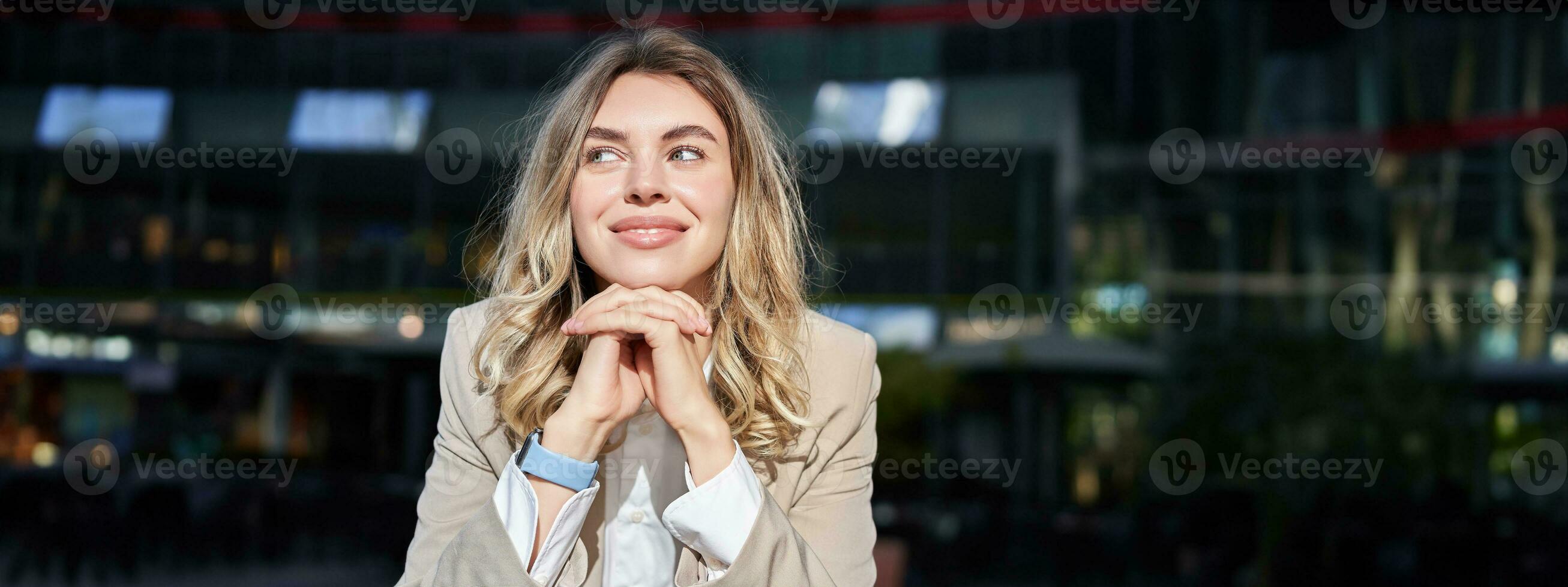 Beautiful corporate woman, looks dreamy and smiles, stands outside on street, leans her head on hands, daydreaming, resting on break after office work photo