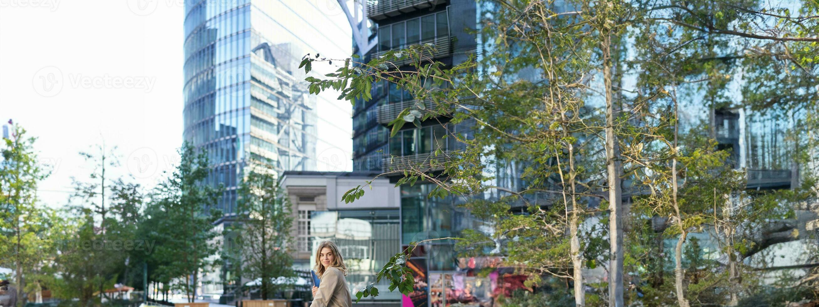 Successful businesswoman in beige suit, standing in city center near sky scrappers and office buildings photo