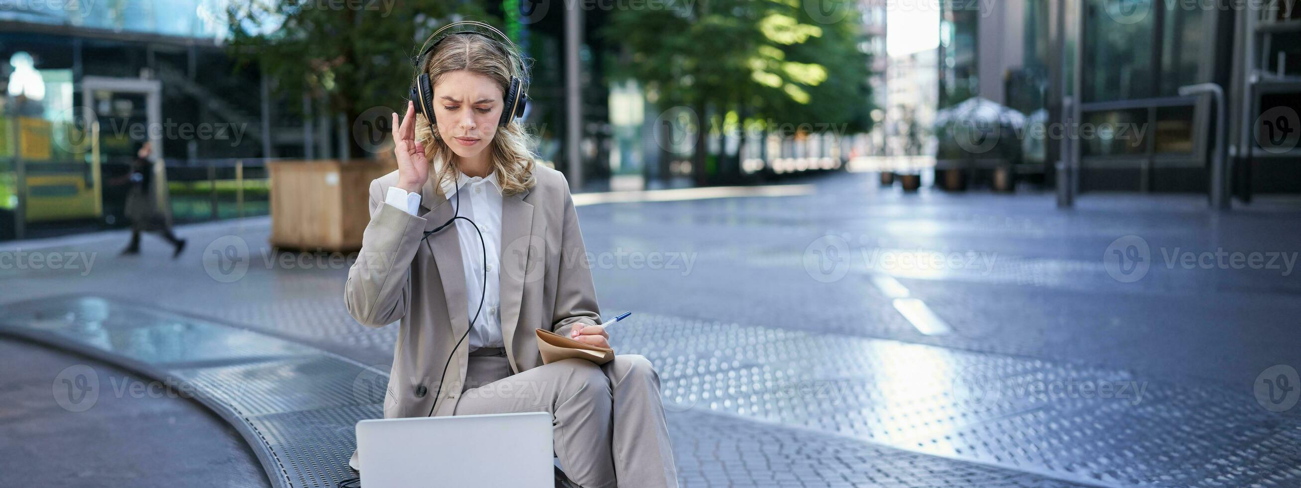 Businesswoman looking perplexed at laptop, taking notes in her notebook, attending online work meeting while sitting on street photo