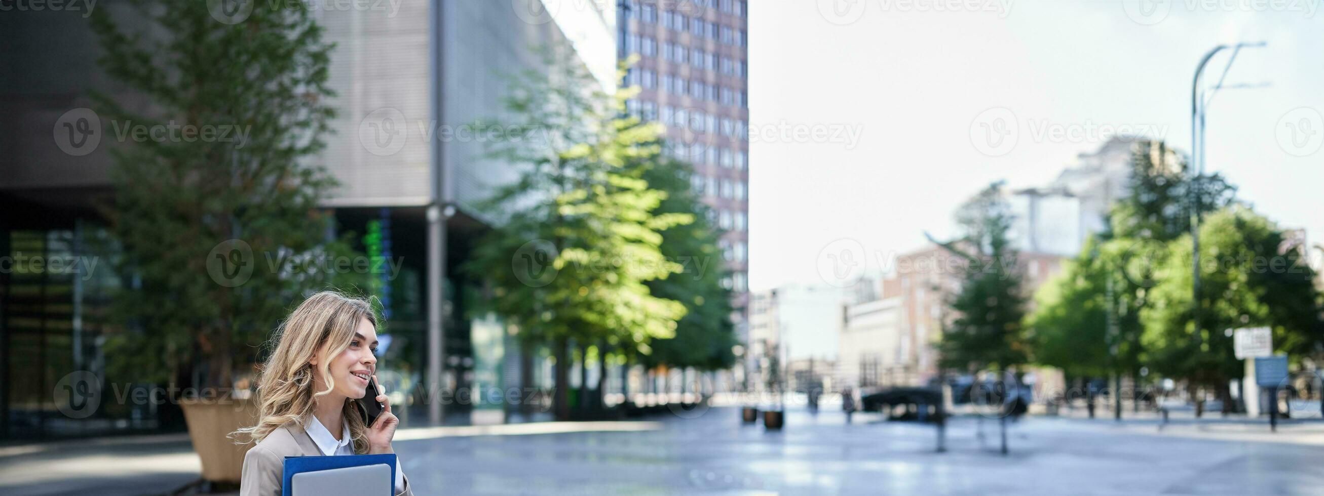 vertical Disparo de joven corporativo mujer con ordenador portátil y trabajo carpetas hablando en móvil teléfono, sentado cerca oficina edificio en ciudad centrar foto