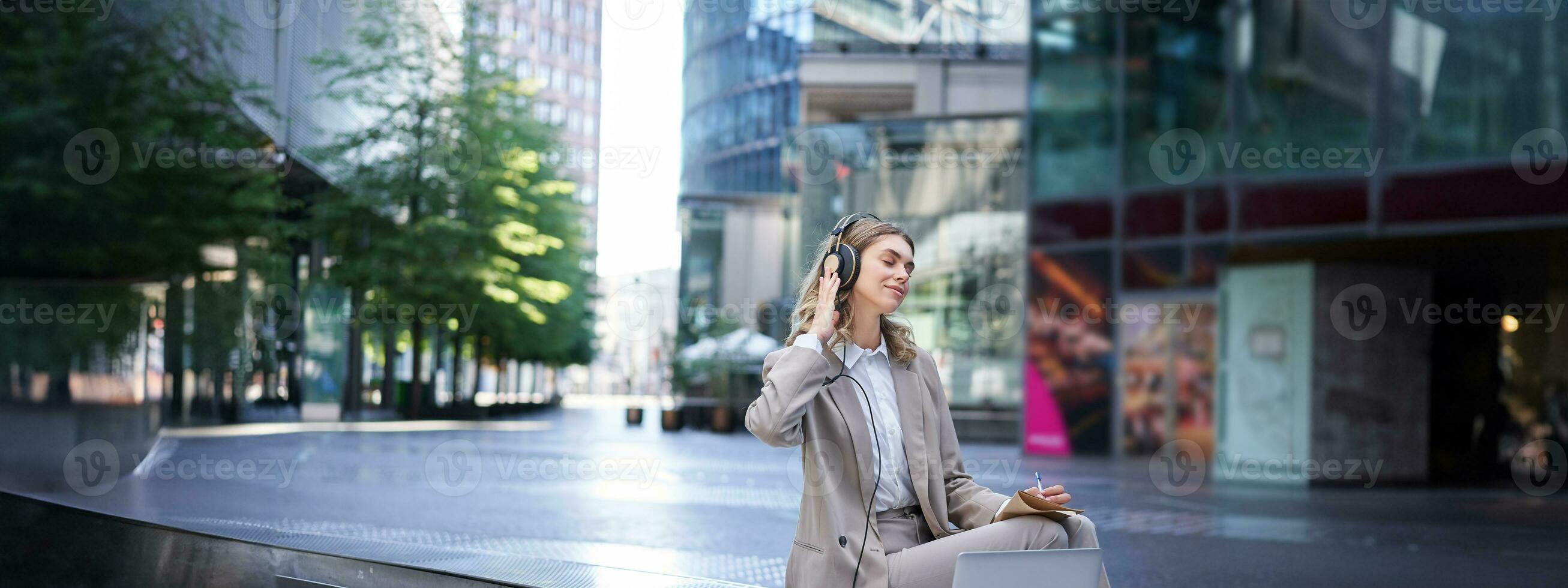 sonriente mujer de negocios relajante, escuchando a música en auriculares desde su computadora portátil, sentado solo en calle de ciudad centro, vistiendo traje foto