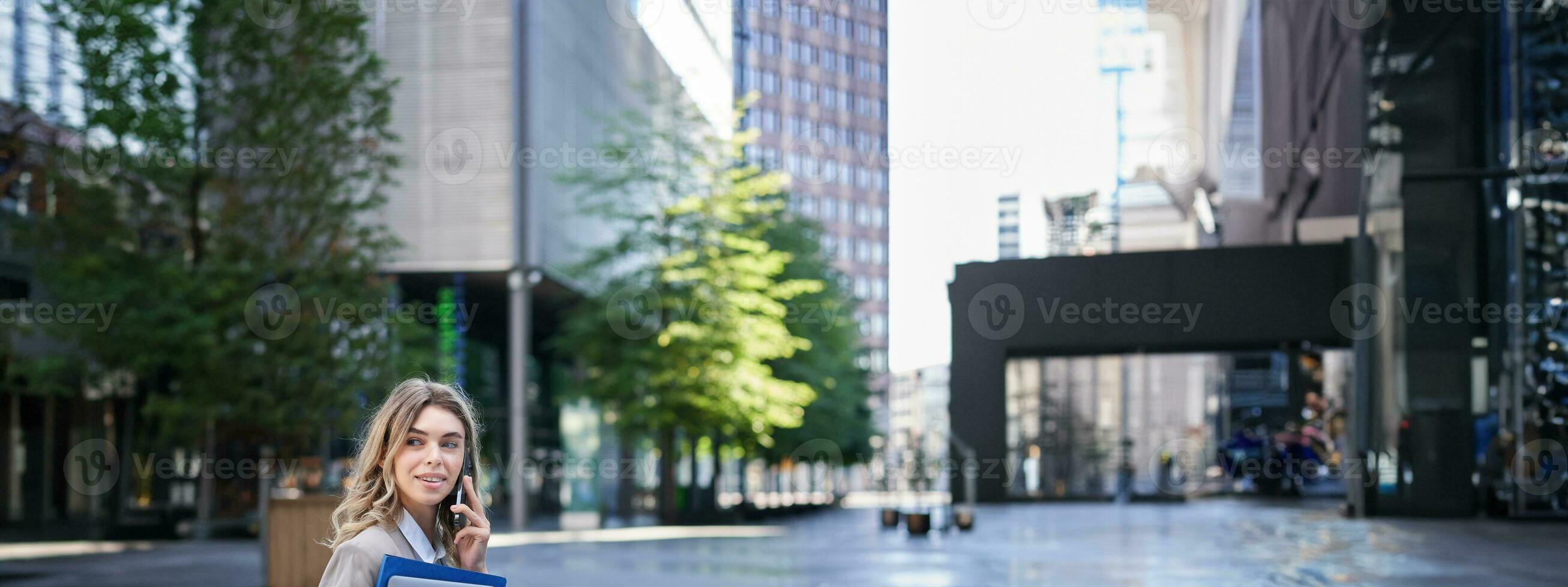 Vertical shot of young woman sitting near office building, holding laptop and folders with work documents, calling someone on mobile phone photo