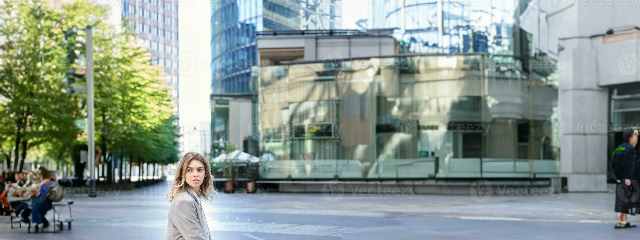 Female corporate worker sitting outdoors near office buildings, turn back and look behind, waiting for coworker on lunch break photo