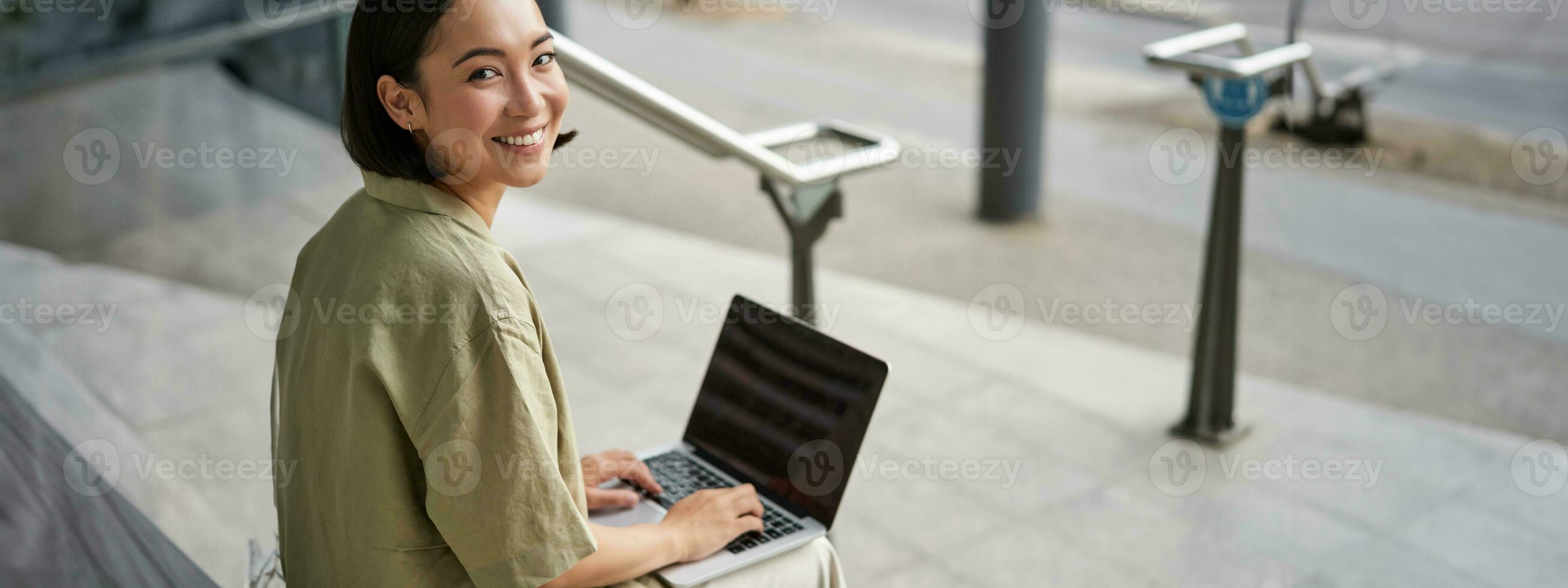 Young smiling woman, asian digital nomad using computer outdoors. Student works on project with laptop, sits on stairs photo