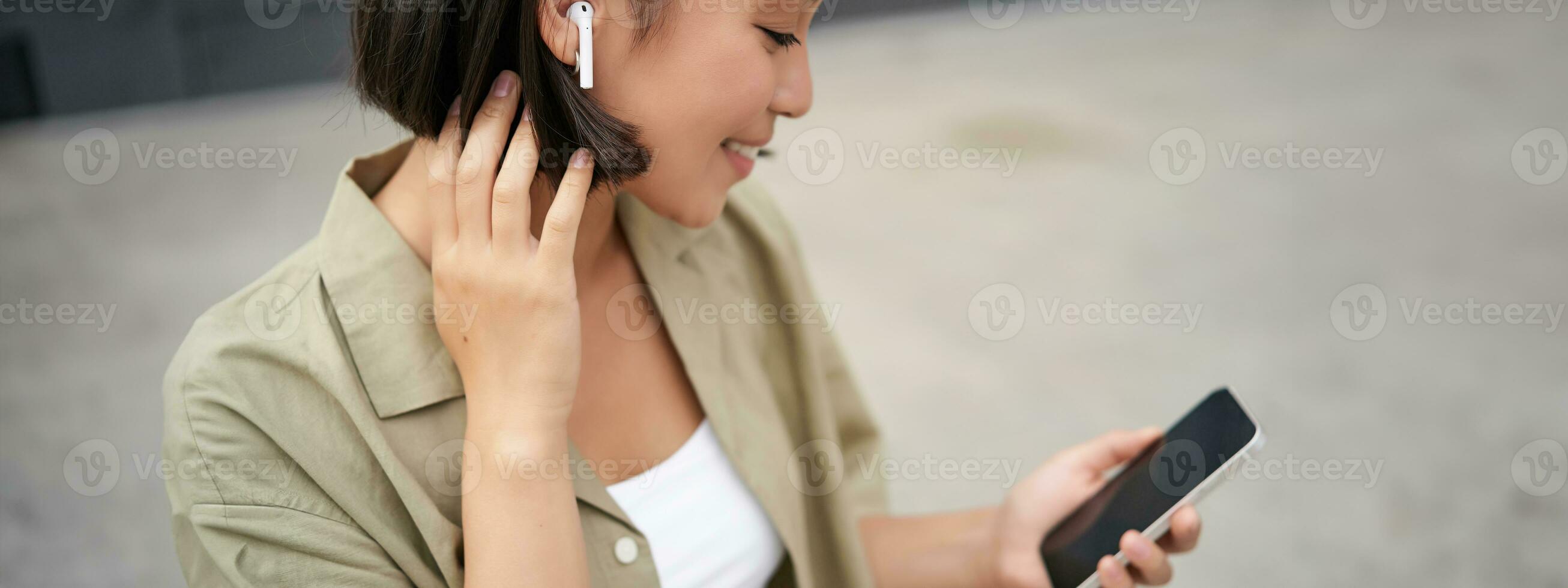 Close up portrait of asian girl, looking at mobile screen, listening music in headphones. Woman with earphones walks on street photo