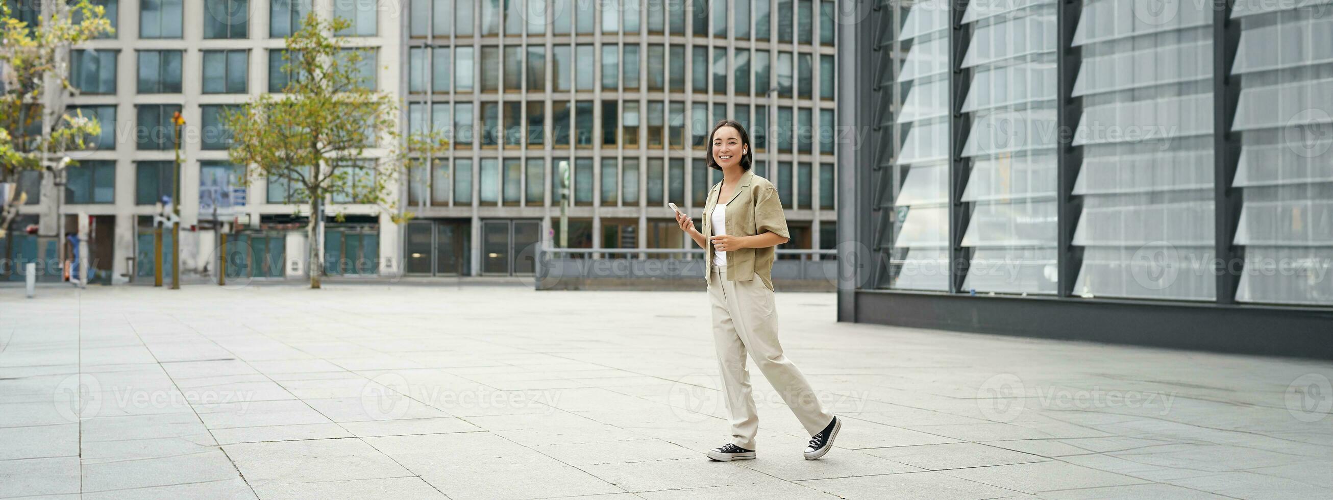 Portrait of beautiful smiling asian woman, walking on street of city centre with smartphone, looking at camera photo