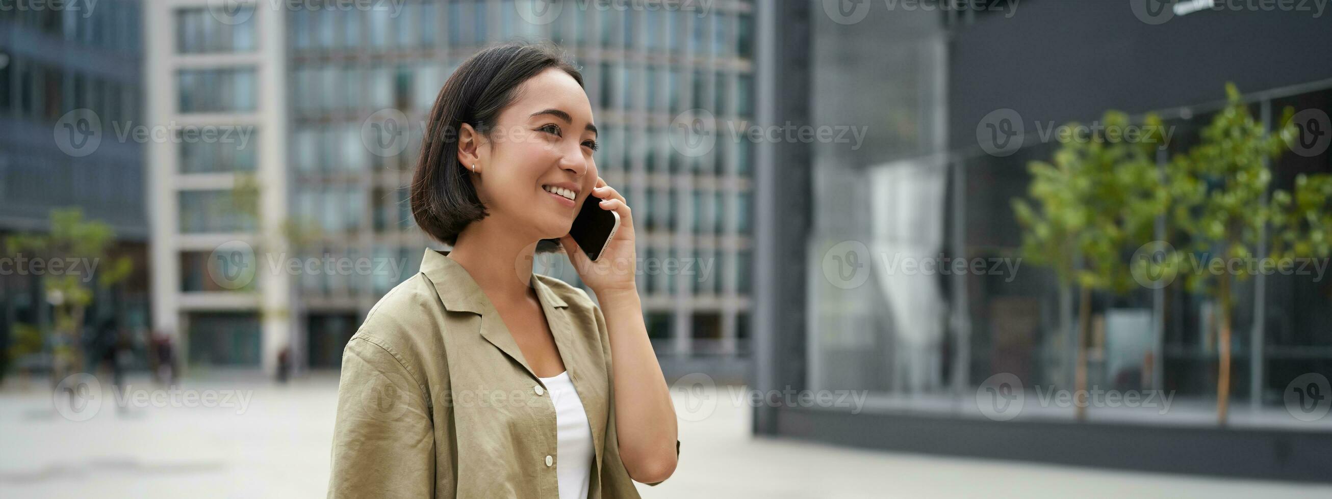 Vertical shot of young asian digital nomad, girl talks on mobile phone and walks on street with laptop. Young woman remote worker going to coworking space photo