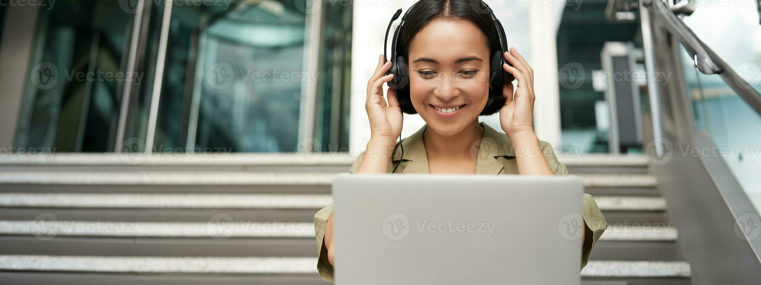 retrato de joven asiático mujer sentado con ordenador portátil y auriculares, acecho video, lo hace en línea curso en computadora, sentado en escalera al aire libre foto
