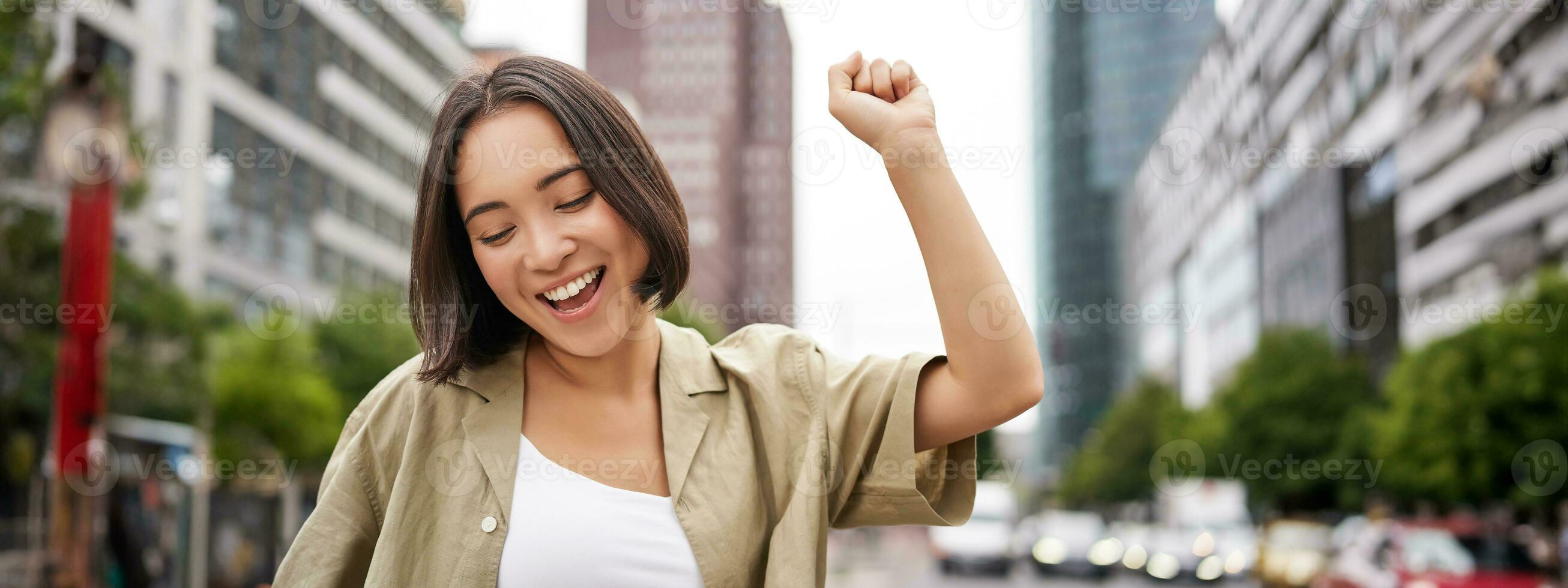 Happy people. Young smiling asian woman dancing in city, raising hands up in city, triumphing, celebrating victory photo