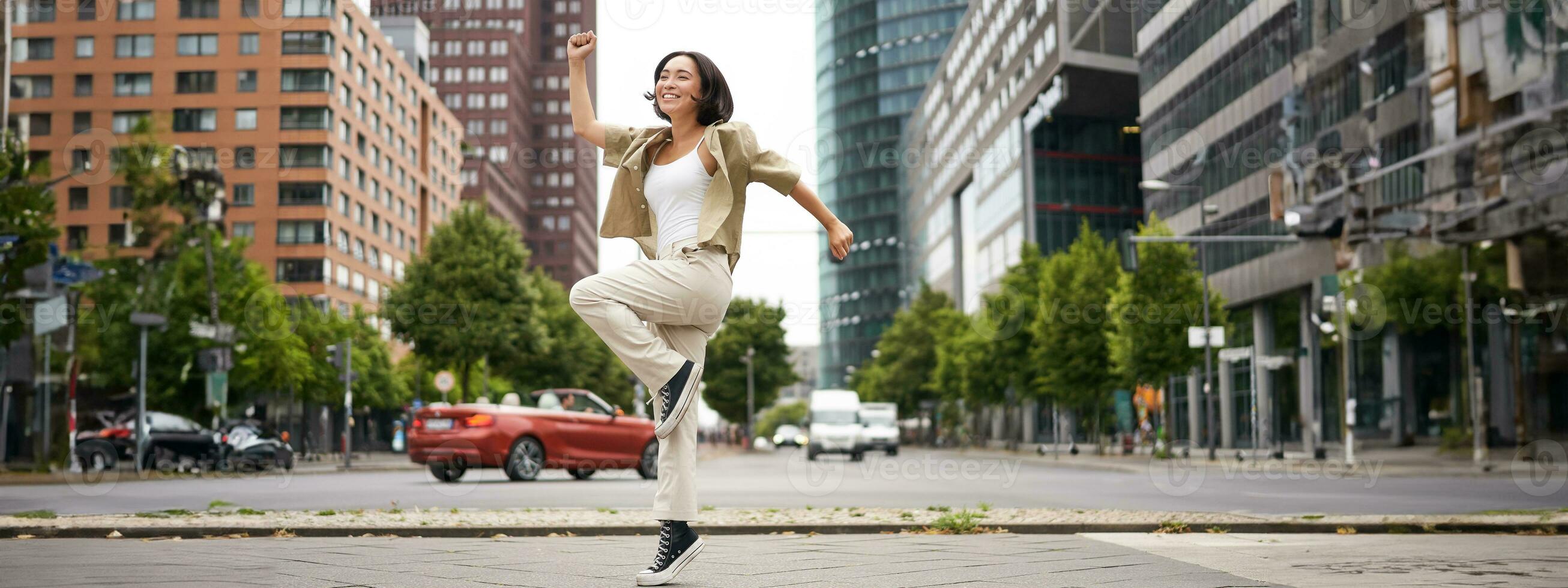 Portrait of asian happy girl jumping and dancing in city centre, posing on streets, express joy and excitement photo