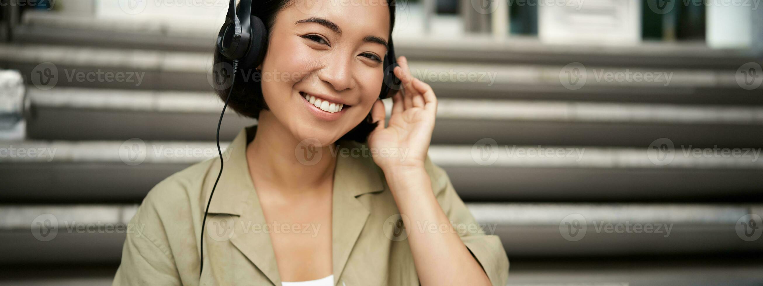 Happy asian girl listens to music in headphones, smiles, sits outdoors on stairs photo