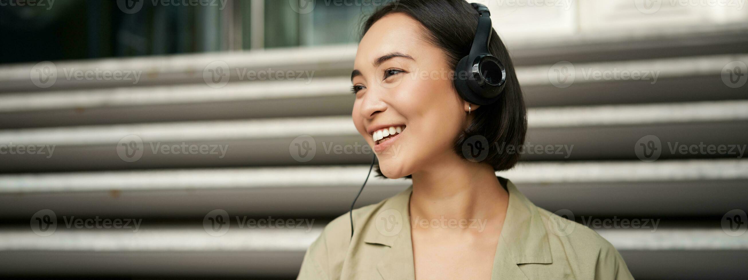 sonriente asiático chica, reír, escuchando música en auriculares, sentado al aire libre. uni estudiante disfrutando gratis hora foto