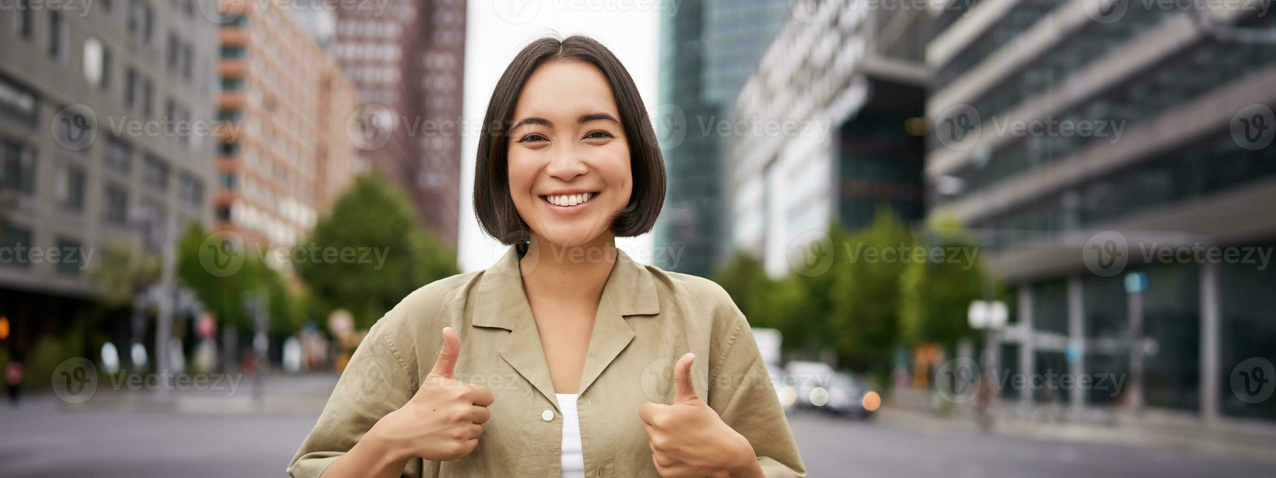 Enthusiastic asian woman , shows thumbs up in approval, looking upbeat, say yes, approves and agrees, stands on street photo