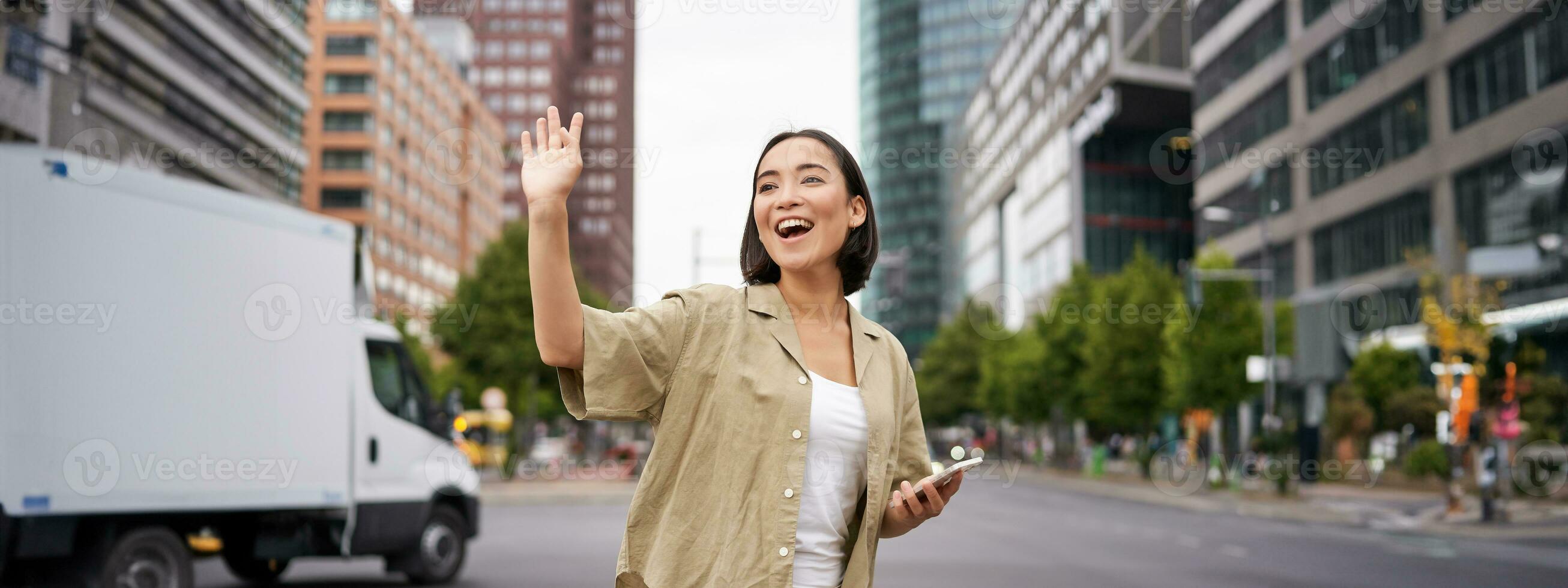 Happy asian girl passing by friend and waving at them on street, saying hello while walking in city, holding smartphone photo