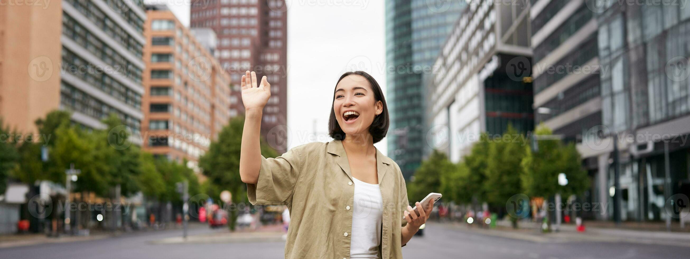 Happy asian girl passing by friend and waving at them on street, saying hello while walking in city, holding smartphone photo