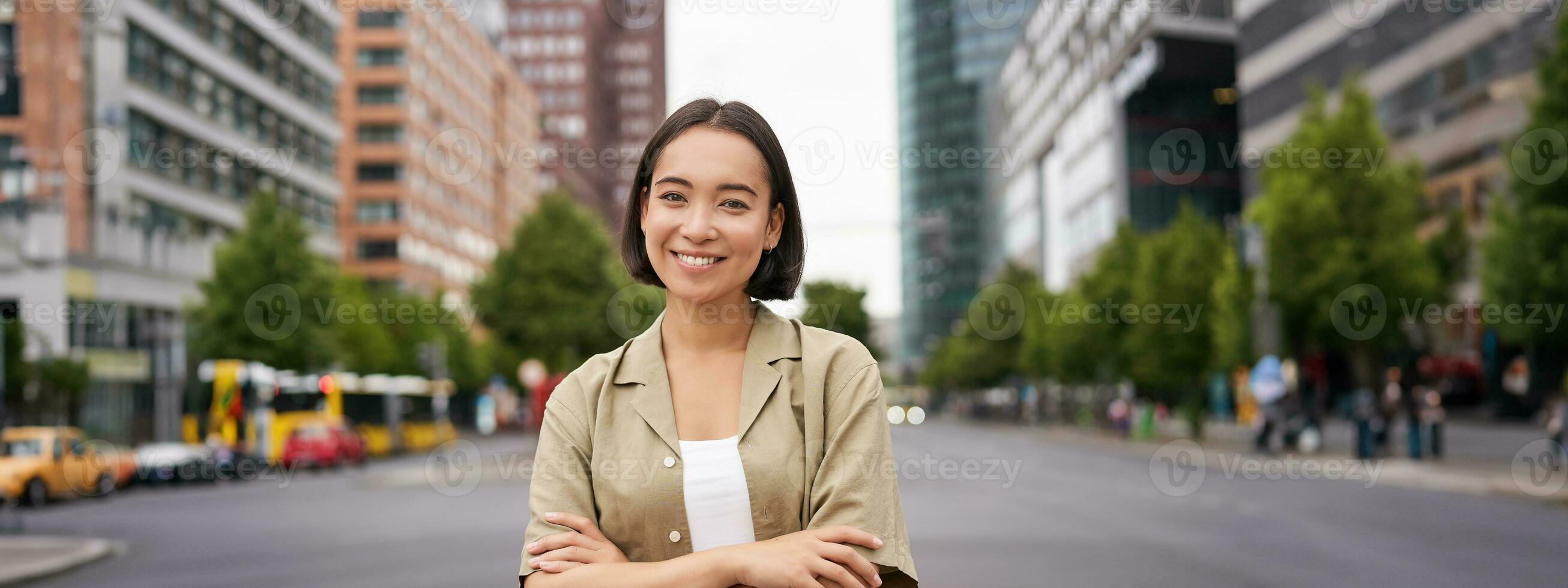 Urban people. Young happy asian girl cross arms on chest, posing on busy city street, smiling with confidence at camera photo