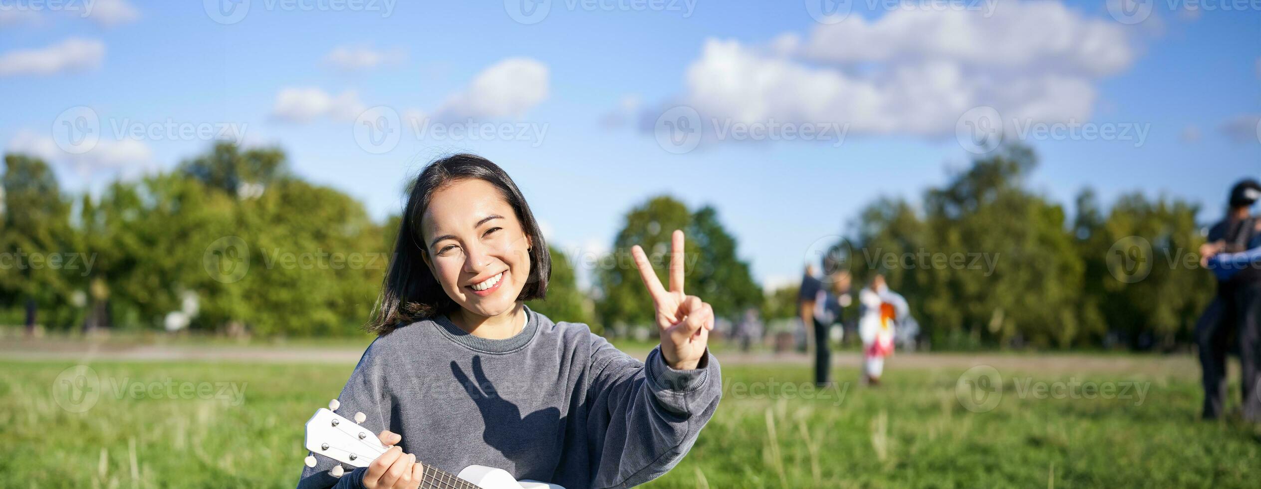 Vertical shot of positive asian girl shows peace sign, plays ukulele in park, rests and enjoys the day. Copy space photo