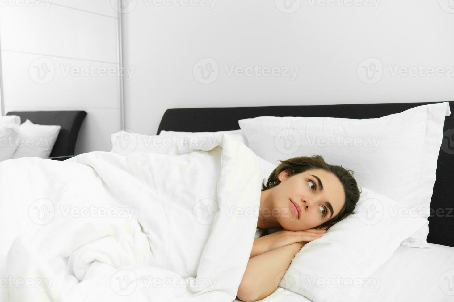 Portrait of dreamy young woman, getting ready for night sleep, lying in bed, covered in white linen sheets, looking aside, waking up in morning photo