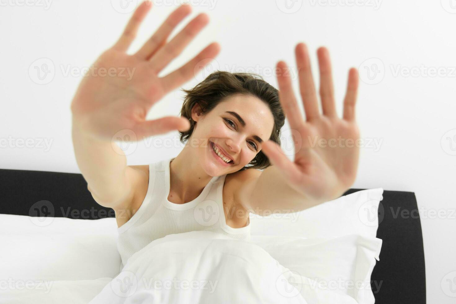 Portrait of sleepy, happy young woman, wakes up in her bed, stretching out hands, hiding face from camera, smiling and laughing photo