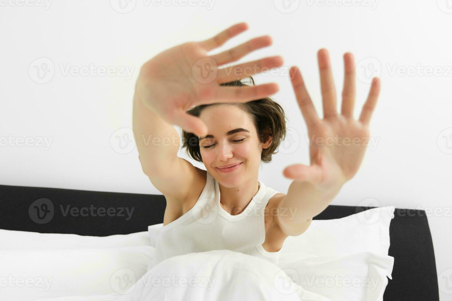 Close up portrait of carefree, beautiful young woman stretching her arms and smiling with dreamy, closed eyes, waking up in morning after good night sleep photo