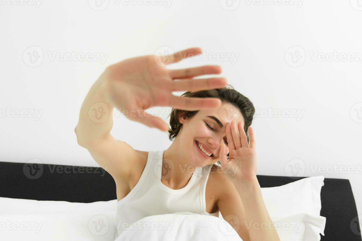 Portrait of sleepy, happy young woman, wakes up in her bed, stretching out hands, hiding face from camera, smiling and laughing photo
