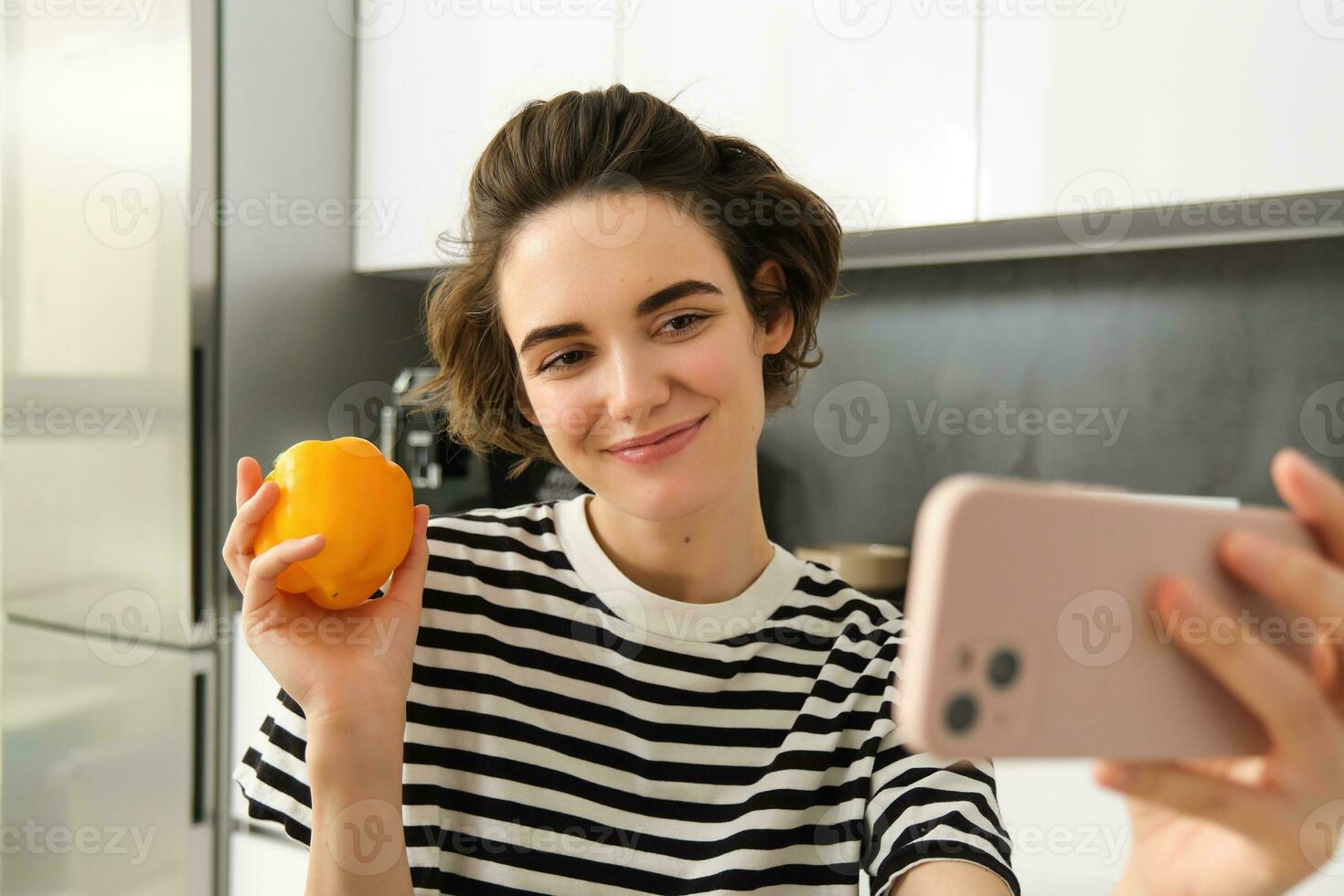 Smiling, beautiful young woman taking selfie with fresh yellow pepper, food blogger taking selfie with vegetables in the kitchen, vegan making a meal and recording video for social media photo