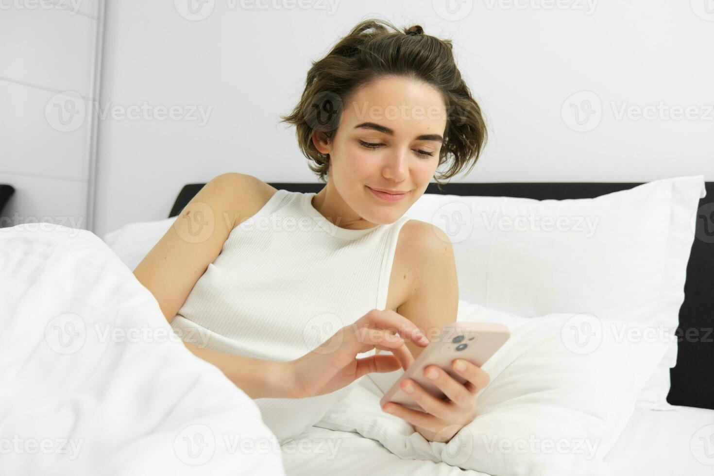 Image of female model in bed, looking at smartphone, sending a message, turns on alarm clock on mobile phone, goes to sleep in her bedroom photo