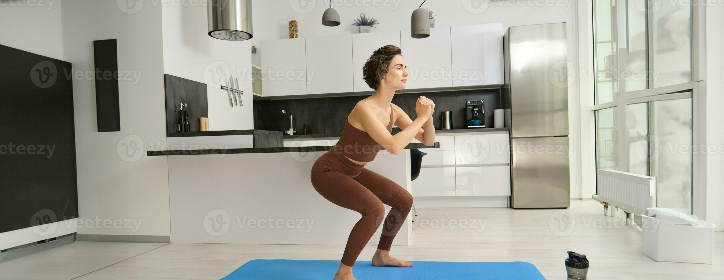 gimnasio a hogar. joven mujer haciendo sentadillas en brillante habitación, rutina de ejercicio adentro en ropa de deporte, haciendo ejercicios en caucho yoga estera foto