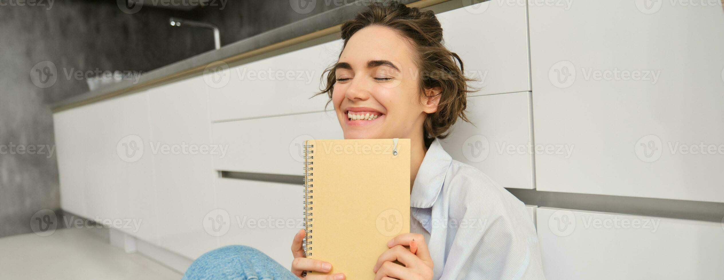 retrato de feliz, despreocupado mujer, escribe en su diario y risas, sostiene su diario, lee alguna cosa en computadora portátil, se sienta en piso adentro foto