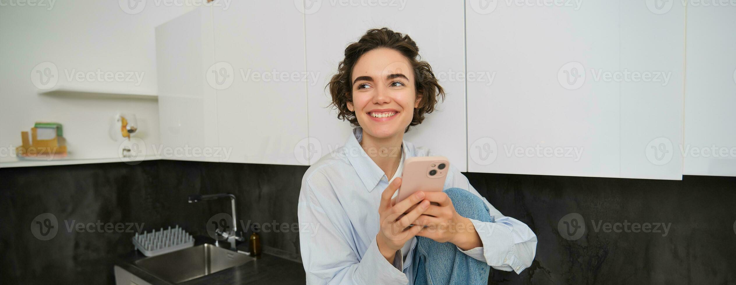 Smiling brunette girl sitting with smartphone at home, orders takeaway from mobile phone app, using online shopping application photo