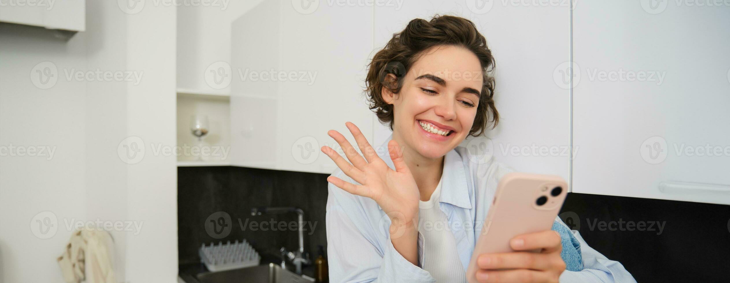 retrato de simpático sonriente morena mujer, sentado a hogar en cocina, mirando a su teléfono inteligente cámara, vídeo chats y olas a móvil teléfono, diciendo Hola a alguien foto