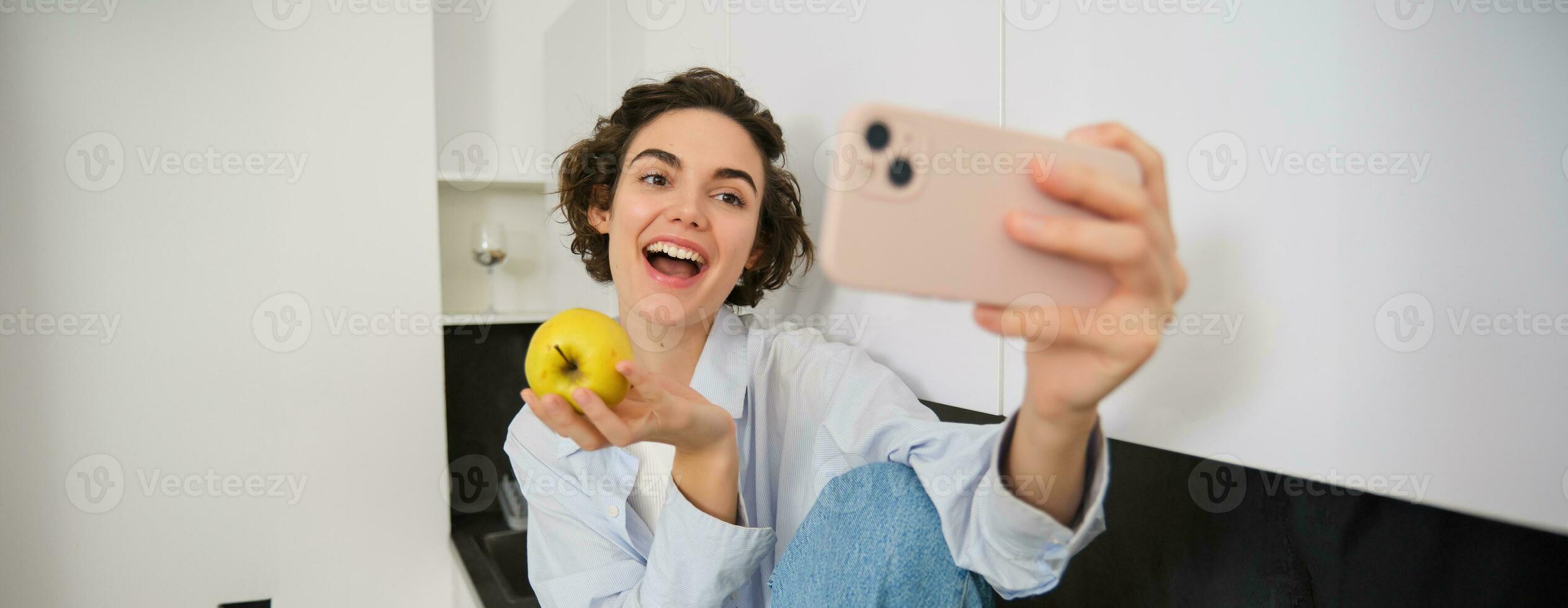 Beautiful woman eating an apple and recording herself on video. Cute girl with fruit, posing for photo, taking selfie in kitchen in happy mood photo