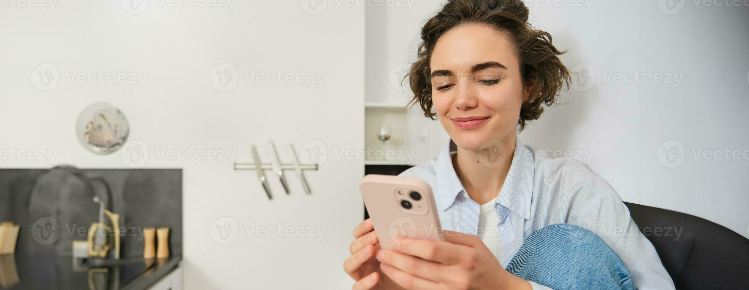 Smiling brunette girl sitting with smartphone at home, orders takeaway from mobile phone app, using online shopping application photo