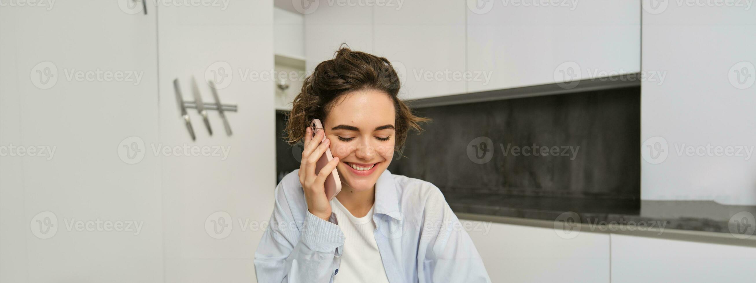 Portrait of smiling young woman talking on mobile phone, laughing, looking down, sitting at home in kitchen photo