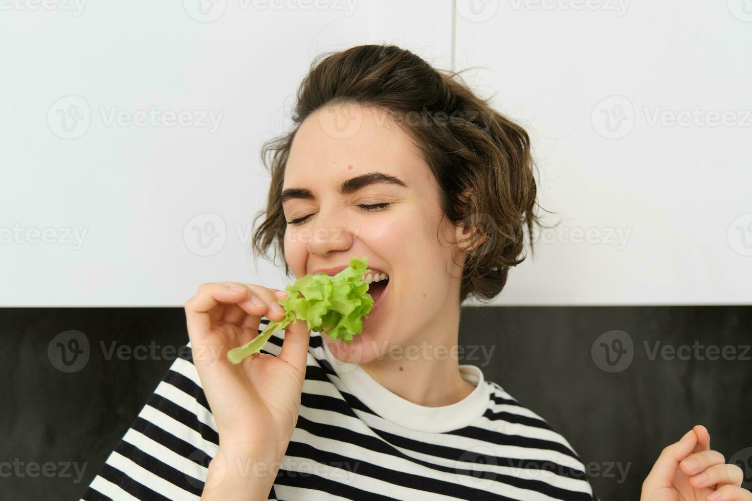 Close up of funny cute woman, vegetarian eating lettuce leaf and smiling, concept of healthy diet, girl likes vegetables, standing in the kitchen photo