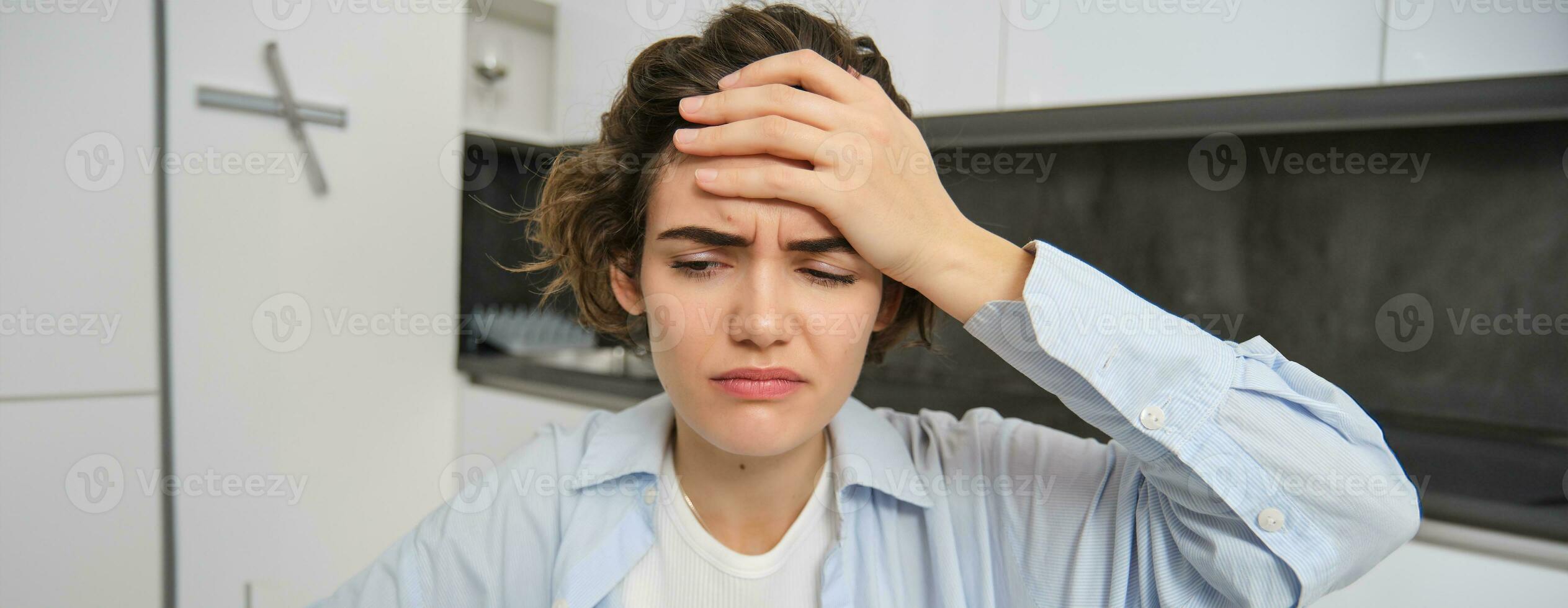 Portrait of woman with headache, sits in kitchen, touches head and grimaces, has painful migraine photo