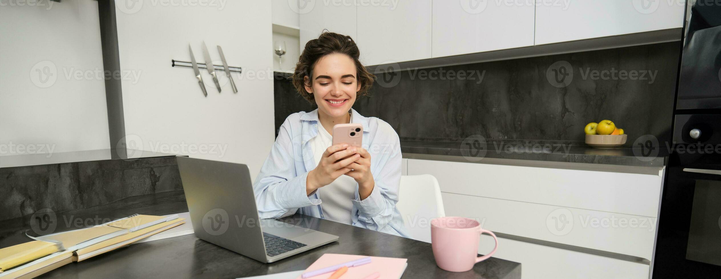 Portrait of brunette woman chats on mobile phone, uses smartphone app from home, orders delivery while sits in kitchen photo
