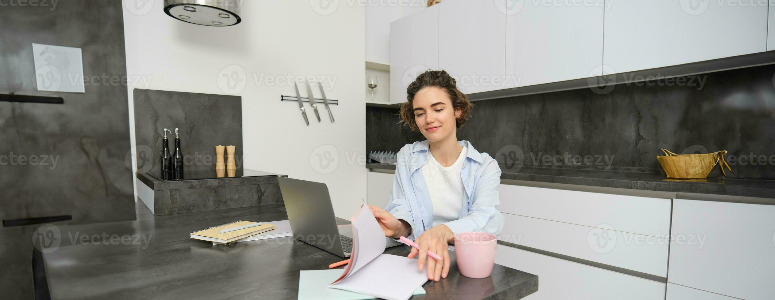 joven determinado mujer, voltea mediante paginas en su libro de trabajo, estudios en remoto, en línea aprendizaje desde hogar, se sienta en frente de ordenador portátil foto