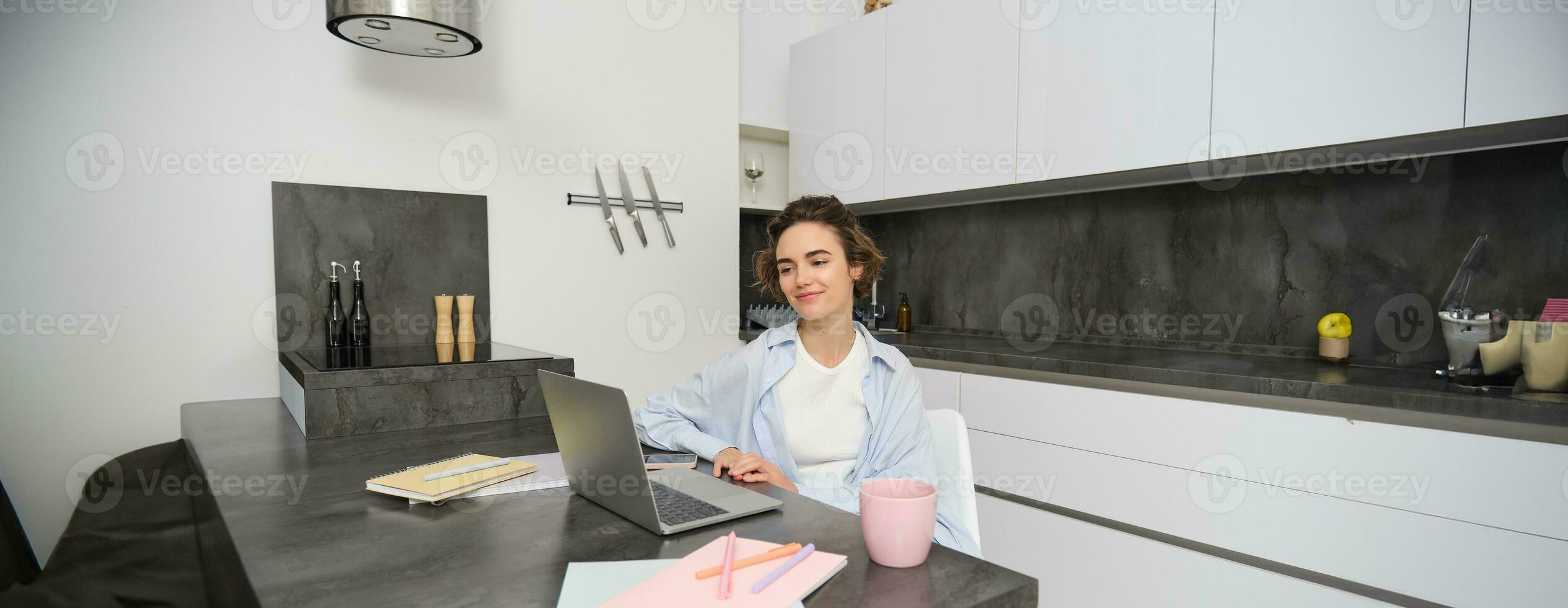 Portrait of young beautiful woman, studies at home, flips her workbook, works from home, uses laptop to learn on remote, makes notes during webinar photo