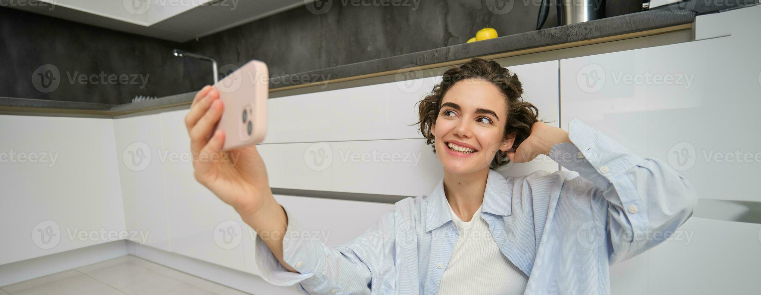 Portrait of young woman sits on kitchen floor with telephone, takes selfie on smartphone with app filters, poses for photo on mobile phone