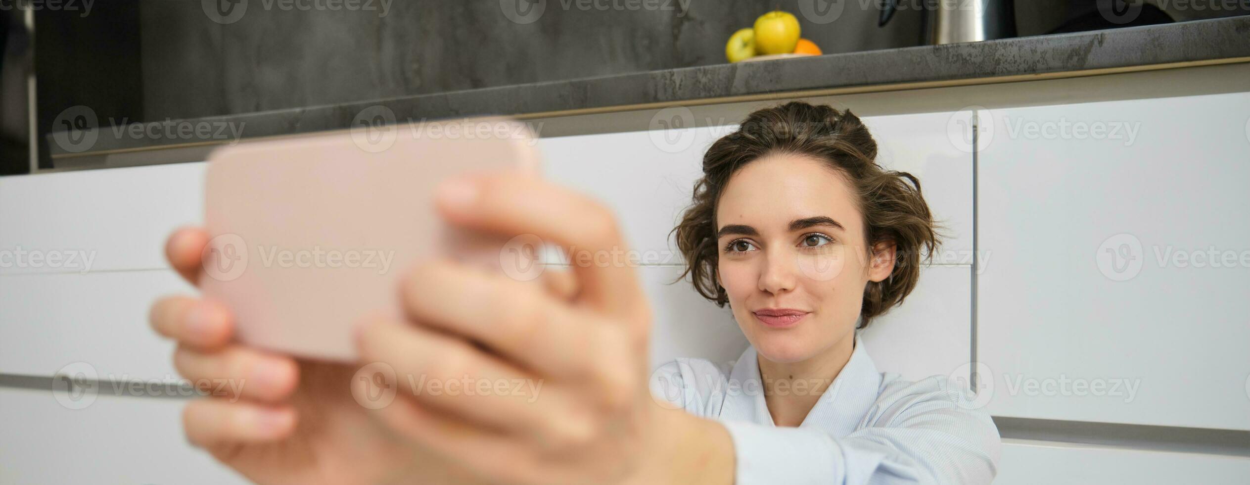 Close up portrait of candid young woman, takes selfie on smartphone, using mobile phone photo app with filters, sits on kitchen floor at home