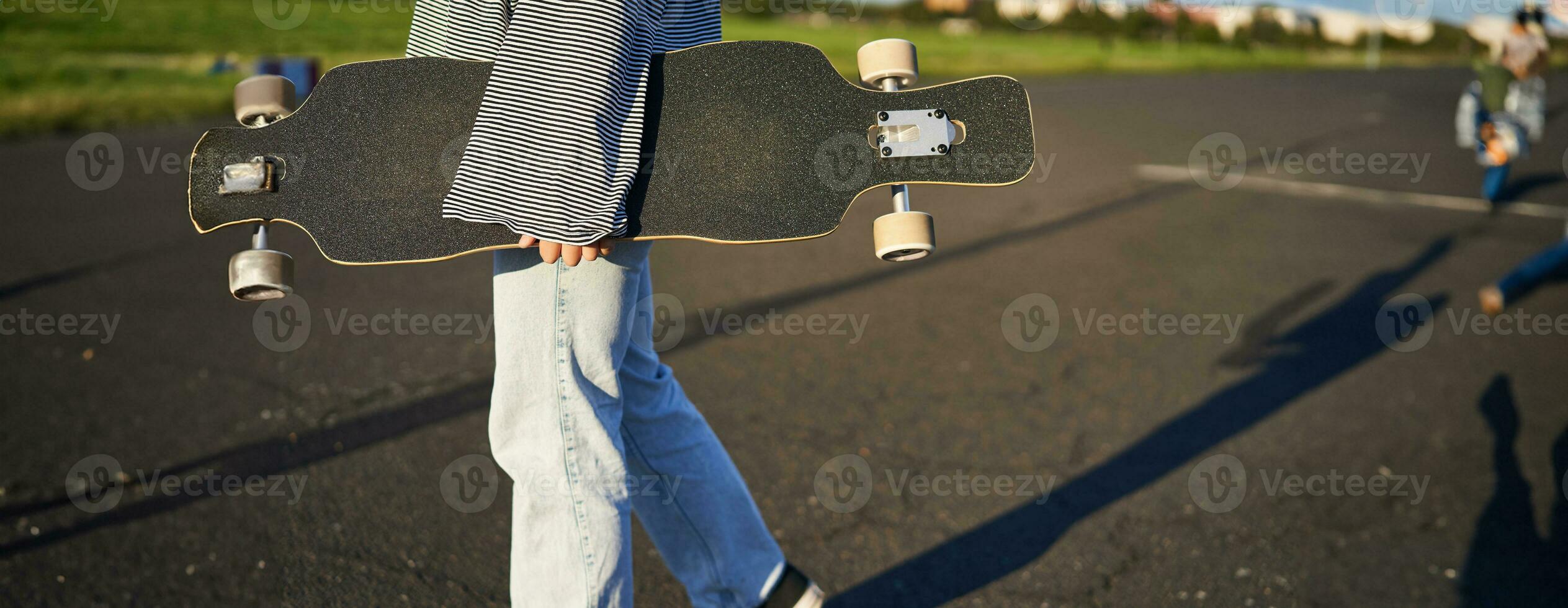 Cropped shot of teen skater girl, hands holding longboard, walking with skateboard on concrete road photo