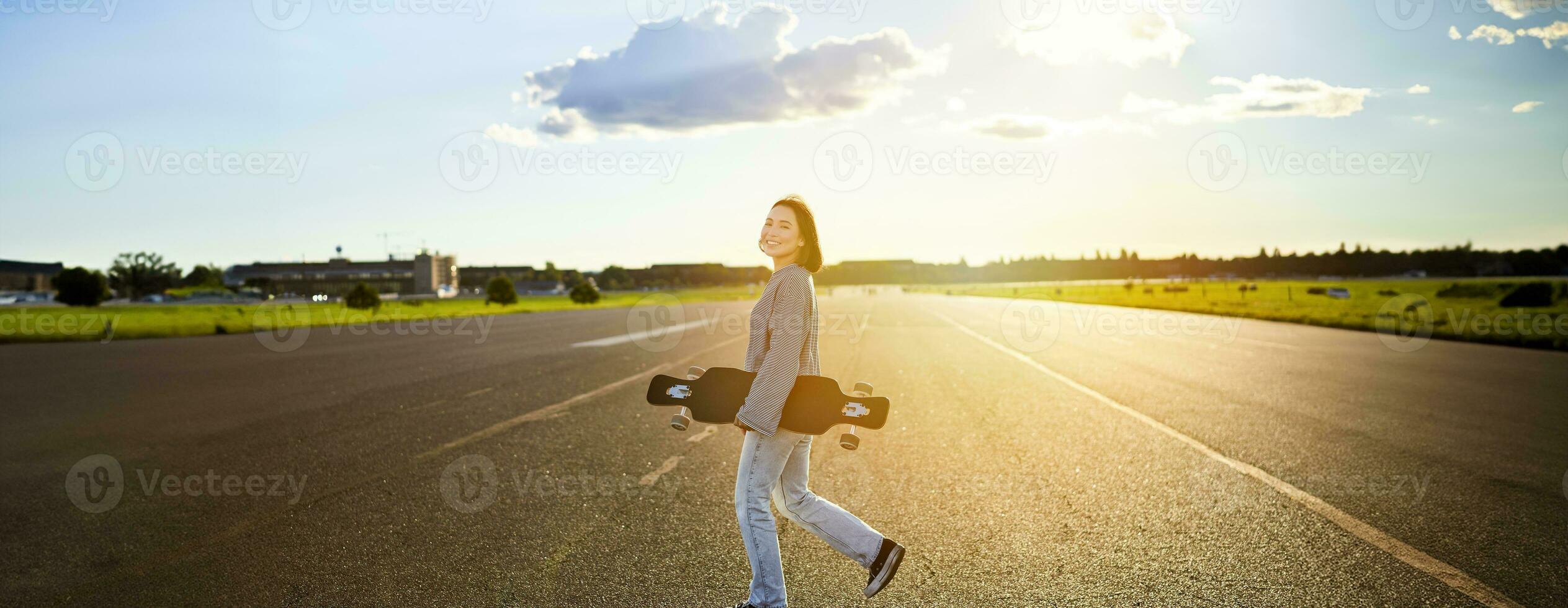 Asian girl with skateboard standing on road during sunset. Skater posing with her long board, cruiser deck during training photo