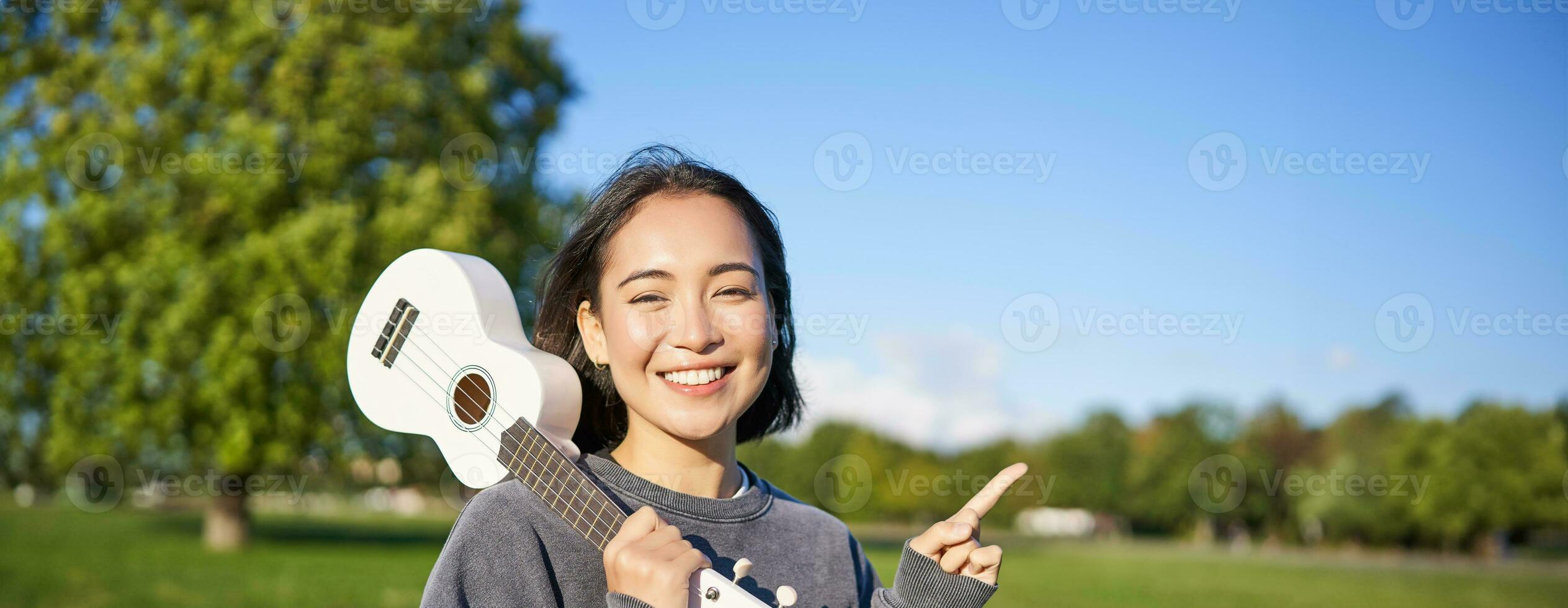 Portrait of asian smiling girl, holding ukulele over shoulder, pointing finger at copy space, banner or logo photo