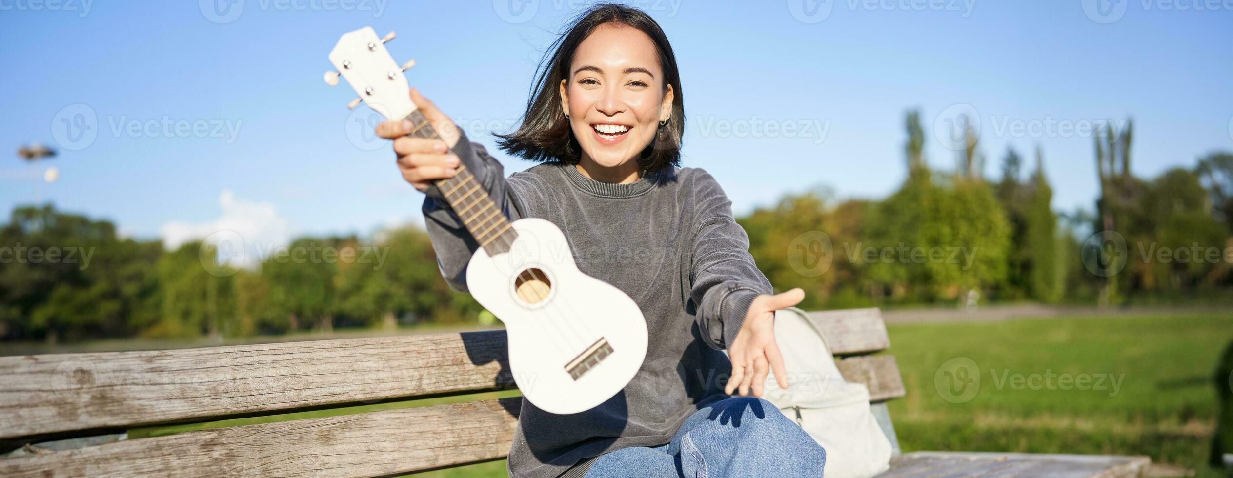 contento asiático niña muestra ukelele, demuestra su nuevo musical instrumento, se sienta en banco en parque y obras de teatro música foto