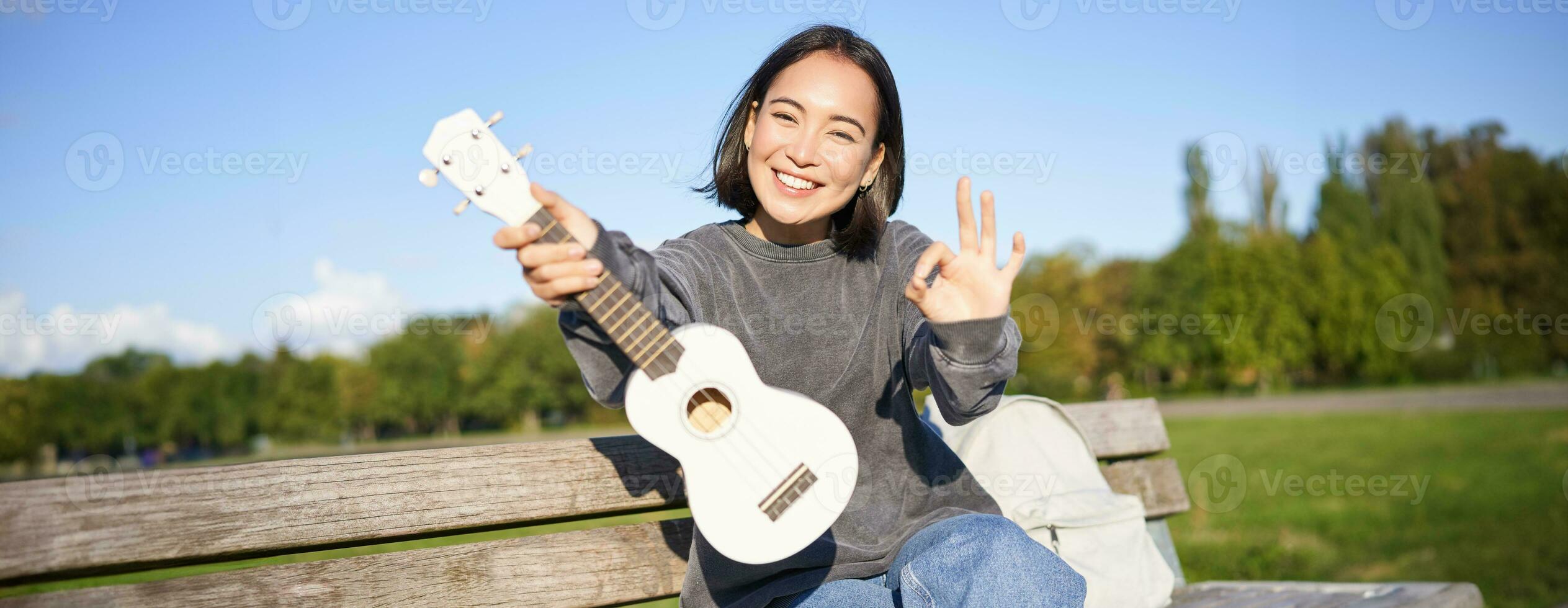 linda sonriente niña muestra Okay firmar y su nuevo ukelele, se sienta en banco en parque, recomienda musical instrumento foto