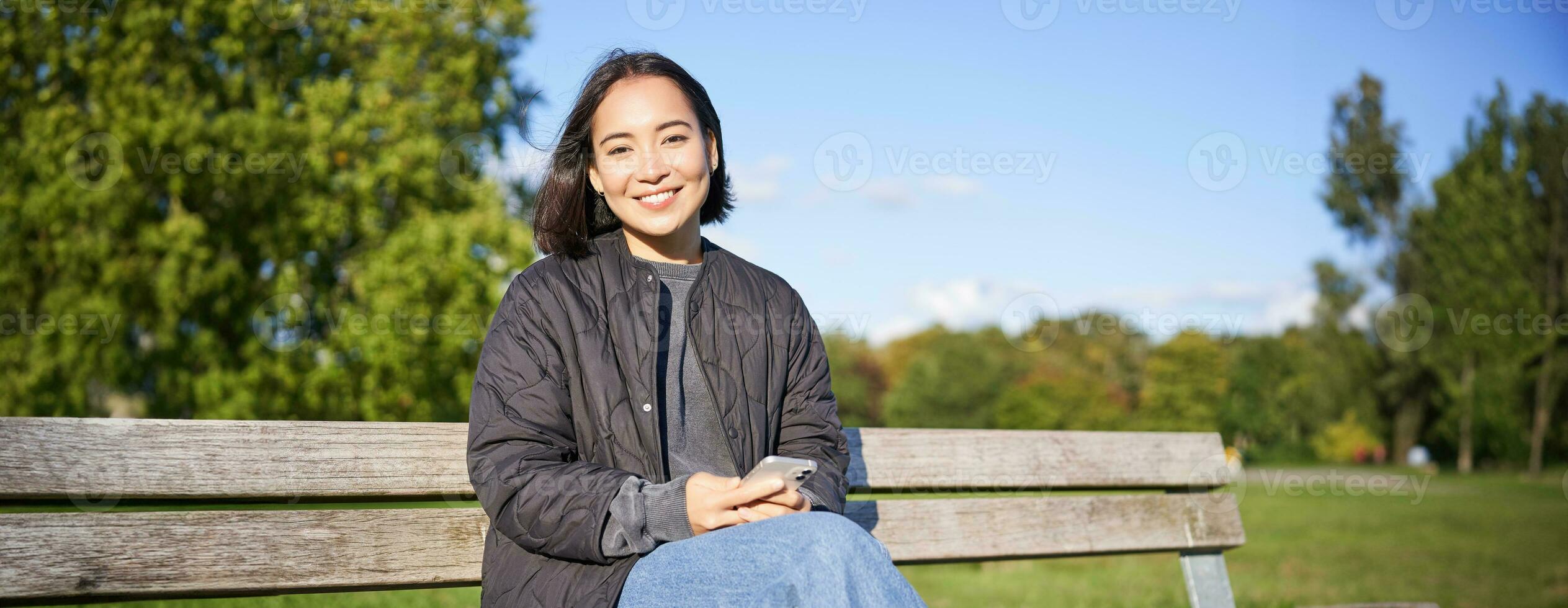 Technology and people. Young woman using smartphone, sitting outdoors on bench in park, looking at smartphone, sending message or networking photo